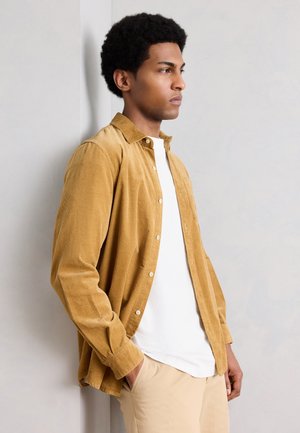 Young man with afro hairstyle leaning against a wall, wearing a mustard yellow corduroy shirt, white t-shirt, and beige pants.
