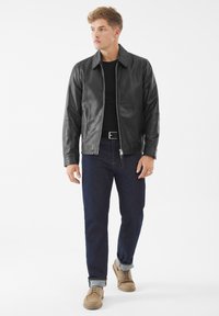 Joven de pie, con chaqueta de cuero negra, camisa negra, jeans oscuros con puños y zapatos beiges con cordones, contra un fondo blanco.