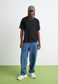 T-shirt en coton noir, jean en denim bleu ample, casquette de baseball grise et baskets blanches avec des accents roses. Tenue simple et décontractée sur un fond uni.