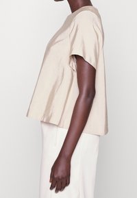 Beige värinen, lyhythihainen yläosa, joka on valmistettu sileästä, hieman kiiltävästä kankaasta. Löysä istuvuus, pyöreä kaula-aukko ja lyhyt, vartalonmyötäinen siluetti.