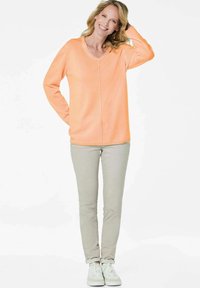 Pullover in sanftem, hellorange Farbton mit V-Ausschnitt und langen Ärmeln. Kombiniert mit hellbeigen Hosen und weißen Sneakers für einen lässigen Look.