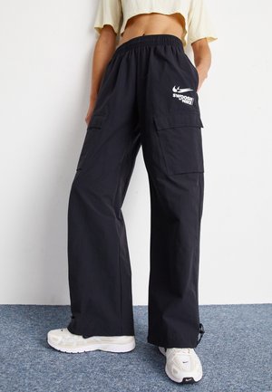 Personne portant un pantalon cargo noir à jambes larges avec le logo Nike Swoosh et des baskets blanches, debout sur un tapis bleu contre un mur blanc.