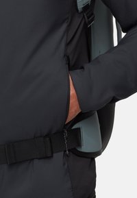 Veste noire isolée avec une poche latérale et des bretelles ajustables. Texture lisse avec un accent gris clair contrastant près de l'épaule.