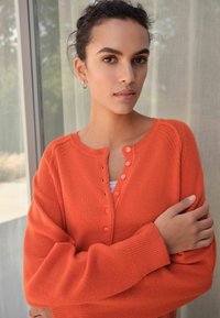 Oranger gestrickter Cardigan mit Knöpfen, runder Halsauschnitt und gerippten Bündchen. Weicher Stoff und lockerer Schnitt.