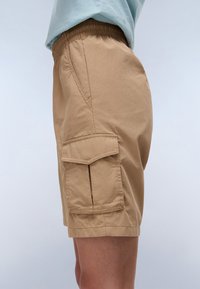 Shorts cargo beiges avec une taille élastique, présentant des poches latérales, une texture cousue et une coupe légèrement ample, affichés de côté.