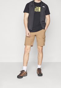Pánský outdoorový outfit skládající se z černé bezrukávové vesty, černého trička se žlutým logem, béžových cargo šortek a hnědých turistických bot s bílými ponožkami.
