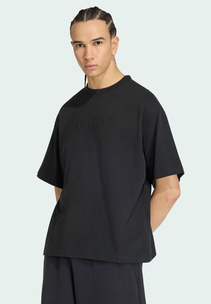 Ung mand med flettet hår iført løs sort Adidas T-shirt og matchende sorte shorts mod en ensfarvet lys baggrund.