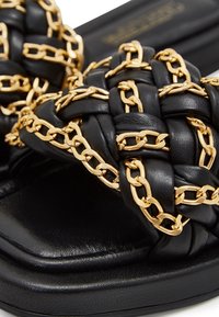 Sandale en cuir noir avec une bride tissée ornée de détails en chaîne dorée et un design à bout carré.