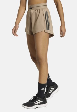 Persona con pantalones cortos deportivos beige con rayas negras, calcetines negros Adidas y zapatillas negras con suelas y rayas blancas.