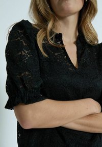 Czarna koronkowa bluzka z bufiastymi rękawami, dekoltem w kształcie litery V z przodu oraz kwiatowymi wzorami na całej powierzchni. Teksturowane wykończenie dodaje głębi i detali.