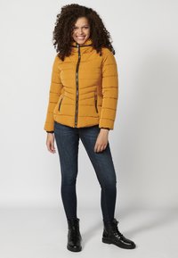 Senapsgul pufferjacka med hög krage, quiltat mönster, svart dragkedja och sidofickor, kombinerat med mörka jeans och svarta stövlar.