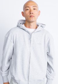 Sivá mikina na zips s mäkkou textúrou, s kapucňou na šnúrky a rebrovanými manžetami. Jednoduchý dizajn s minimalistickým brandingom na hrudi.