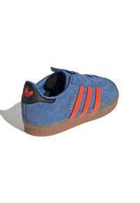 Blå sneakers i mocka med tre oransje striper, svart hælakkent og en gjennomsiktig gummisåle. Modellenavn "Gazelle" vises.