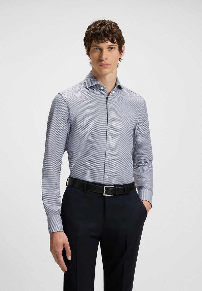 Camicia da uomo button-down a scacchi blu e bianchi, con maniche lunghe, colletto e una texture di tessuto liscia, abbinata a pantaloni scuri.
