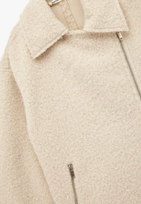 Crème-kleurige fleecejas met een gestructureerd oppervlak, voorzien van een kraag en een ritssluiting aan de voorkant. Zilveren ritsbeslag zichtbaar.