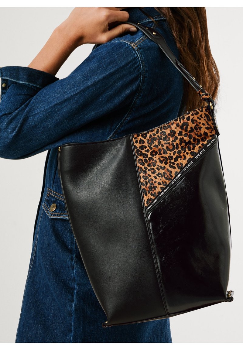 Femme portant une veste en jean foncée et un grand sac à bandoulière en cuir noir avec un panneau imprimé léopard et une bordure de marque.