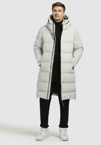 Lys grå pufferjakke med hætte, der har en lang silhuet, tekstureret polstring, lynlåslukning og sidelukninger, båret over et sort outfit.