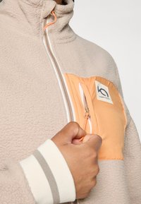 Beige fleecejakke med en frontlomme i orange, featuring lynlås lukning og logo patch. Kraven er høj, og ærmerne har grå striber.