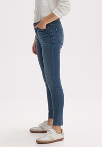 Modré úzké džíny s hladkou texturou, vybavené pětikapsovým střihem a jemným prošíváním. Kombinovány s bílými sportovními teniskami pro neformální vzhled.