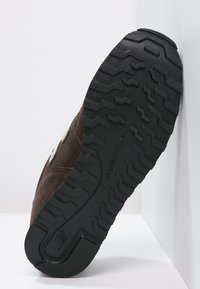 Semelle en suède brun avec une semelle extérieure en caoutchouc noir texturé présentant un motif en zigzag pour une meilleure adhérence. Nom de la marque embossé sur le côté.