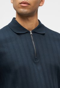 Νεαρός άνδρας που φοράει μπλε ριμπ πουκάμισο πόλο με ασημί ημί-φερμουάρ και στρογγυλεμένο γιακά, μπροστά σε απλό ανοιχτό φόντο.