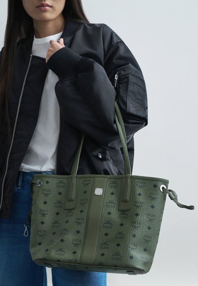 Persoană îmbrăcată într-o jachetă bomber neagră și o cămașă albă, ținând o geantă mare cu model verde și mânere duble.