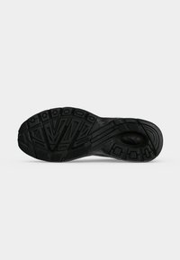 Semelle de chaussure en caoutchouc noir avec motifs géométriques de la bande de roulement et zones de préhension texturées sur un fond uni clair.