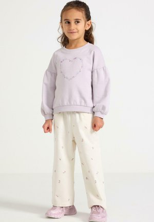 Vaaleanpunainen svetari, jossa on pussihihat ja kirjailtu sydänyksityiskohta, yhdistettynä kermaan värisiin housuihin, joissa on pieniä kukkakuvioita. Vaaleanpunaiset urheilukengät.