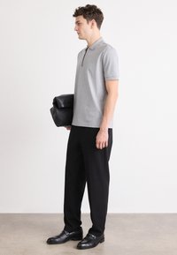 Šedé polo tričko s krátkymi rukávmi a zipsovým výstrihom, kombinované s čiernymi nohavicami a čiernymi topánkami. Drží čiernu koženú tašku.