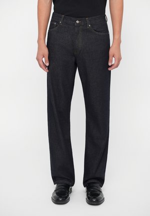 Persona che indossa jeans in denim nero a taglio ampio con bottoni e cerniera frontale, abbinati a scarpe in pelle nere e una maglietta nera.