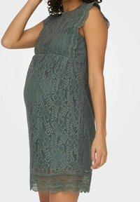 Spitzen-Maternity-Kleid in Dunkelgrün mit gewelltem Ausschnitt und Saum, mit floralen Mustern und lockerer Passform, um einen wachsenden Babybauch zu accommodate.