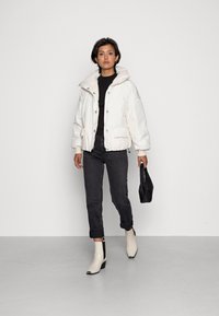 Veste blanche rembourrée avec un col haut, fermeture à boutons et deux poches avant. Portée sur un haut noir, associée à un jean sombre et des bottines claires.