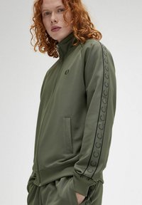 Giacca zip-up verde oliva con colletto alto, due tasche laterali e logo. Presenta un motivo a strisce testurizzate lungo le maniche.