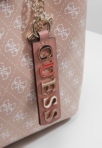 Różowa torebka z ekoskóry z białym wzorem logo, złotym łańcuszkiem oraz brązową metką z napisem "GUESS" w złotych i czerwonych literach.