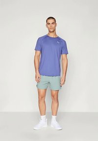 Hombre de pie con camiseta púrpura de Puma, pantalones cortos verde claro, calcetas blancas y zapatillas blancas frente a un fondo claro y liso.