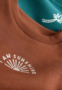 Två tröjor staplade; en brun med texten "I AM SUNSHINE" och solgrafik, den andra teal med vit blommig broderi. Bomullsmaterial, ribbade halslinningar.