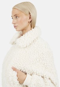 Witte textuur jas met een pluizige uitstraling, brede kraag en ribgebreide manchetten. Bevat zijzakken die vanaf de voorkant toegankelijk zijn.