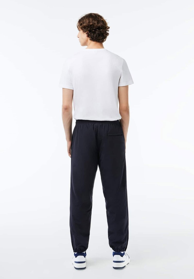 Jeune homme aux cheveux bouclés se tenant de dos, vêtu d'une chemise blanche à manches courtes, d'un pantalon de jogging noir à taille élastique et de baskets blanches.