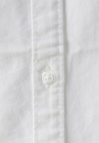 Tessuto di camicia in cotone bianco con un bottone visibile, caratterizzato da una consistenza liscia e una leggera lucentezza. Cucitura lungo il bordo.