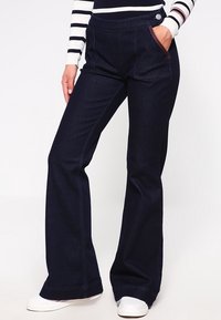 Donkerblauwe flare jeans met een gladde denimtextuur. Voorzien van een brede tailleband, een enkele voorkauw met contrasterende stiksels en een knoopsluiting.