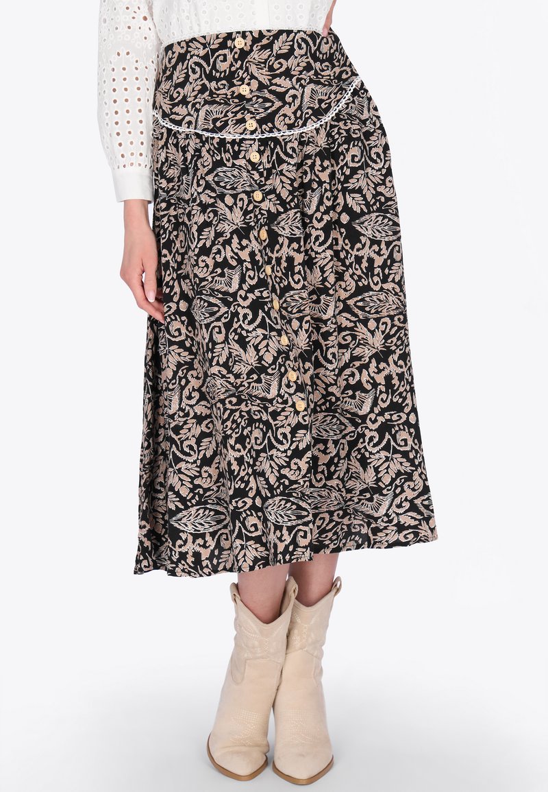Nő fekete midi szoknyában, bézs virágmintákkal, gombos elülső résszel, fehér lyukacsos blúzzal és bézs bokacsizmával.