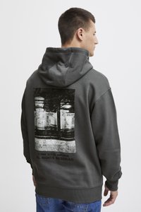 Young man facing sideways wearing dark gray hoodie with black and white graphic and text "© 1999 SOLID ARCHIVE ALL RIGHTS RESERVED" on back.