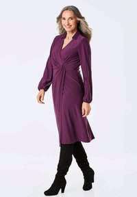 Lila Wickelkleid mit langen Ärmeln, gerafftem Vorderteil und knieumspielendem Saum. Getragen mit schmalen schwarzen Overknee-Stiefeln.
