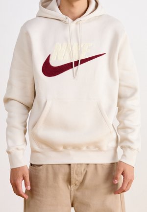 Person iført cream-farvet Nike hoodie med rød swoosh-logo og beige bukser, stående mod en ensfarvet baggrund.