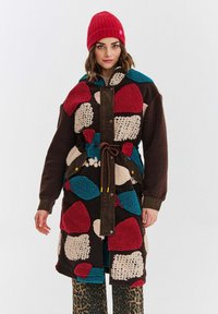 Žena oblečená v farebnom patchwork flísovom kabáte so hnedými rukávmi, červenou pletenou čiapkou a nohavicami s leopardím vzorom na pozadí čistej bielej.