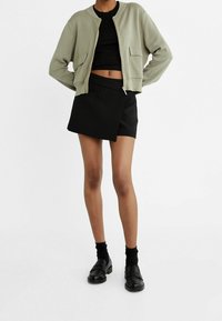 Veste courte olive, haut noir ajusté, jupe portefeuille noire et chaussures noires à finition lisse ; dispose de manches courtes et de poches avant.