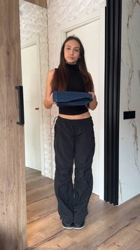 Mladá žena s dlhými vlasmi v čiernom tielku bez rukávov a nohaviciach stojaca vo vnútri, držiaca zložené modré rifle pred bielymi dverami.