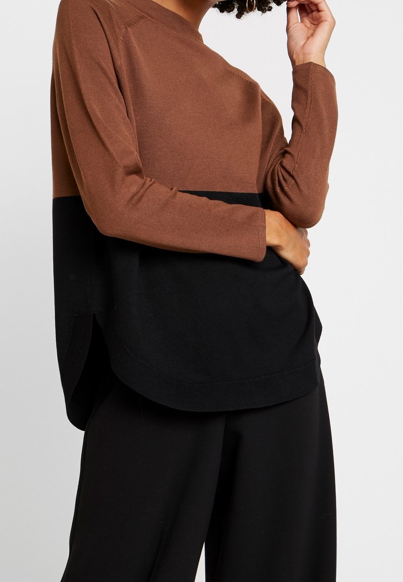 Femme portant un pull bicolore marron et noir à manches longues avec un pantalon noir ample, debout avec une main près du visage.