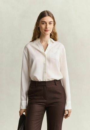 Jonge vrouw met lang haar draagt een wit overhemd met knopen en donkerbruine broek, en houdt een zwart voorwerp in haar linkerhand.