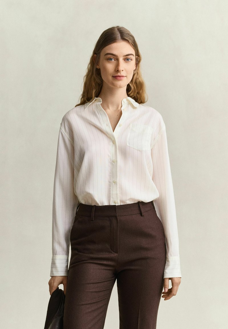 Jeune femme aux longs cheveux portant une chemise blanche boutonnée et un pantalon marron foncé, tenant un objet noir dans sa main gauche.